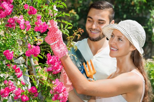 A smiling man and woman are working together in a lush garden, with the woman wearing a wide-brimmed hat and gardening gloves, tending to a vibrant pink flowering shrub. The man stands close by, holding a small garden fork, both focused on the healthy, blooming plant. The garden features a well-maintained lawn with dense, green grass, bordered by flower beds and a hedge, with tall trees visible in the background. The scene depicts outdoor landscaping or gardening activity, highlighting elements such as flowering plants, neatly trimmed hedges, and a lawn area, all bathed in natural daylight with a clear, pleasant weather. This setting reflects typical garden maintenance and landscaping services offered by companies such as Lawn Mowing Palmers Green, catering to residential outdoor spaces near Palmers Green, N13, in North London, which include lawn care, plant maintenance, and shrub trimming.