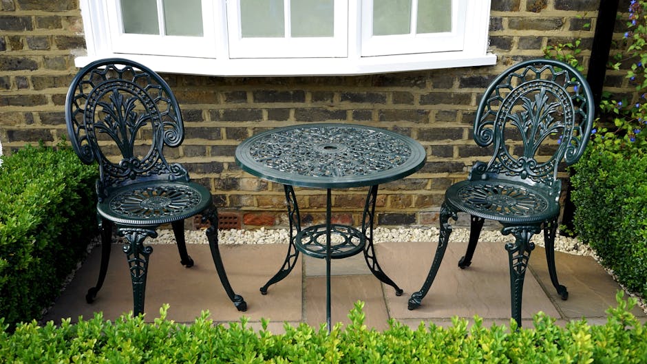 A small outdoor garden area featuring a decorative wrought iron table with an intricate floral pattern on its surface, accompanied by two matching wrought iron chairs with ornate backrests and curved